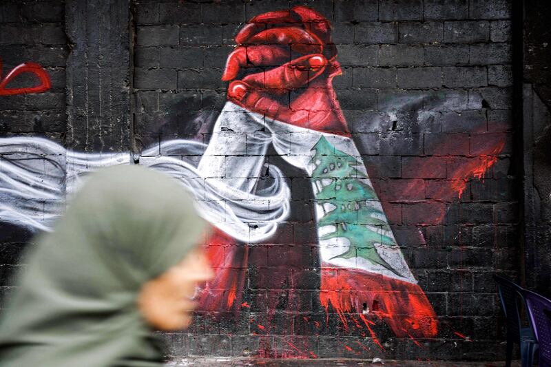 This picture taken on October 24, 2019 in the northern Lebanese city of Tripoli shows a woman passing by graffiti depicting two hands holding each other, both painted in the colours of the Lebanese national flag, painted by Palestinian refugee Ghayath al-Rawbeh, who was originally a Palestinian refugee in Syria and fled with the start of the Syrian crisis, painted on a wall at al-Nour Square in the city centre.  - RESTRICTED TO EDITORIAL USE - MANDATORY MENTION OF THE ARTIST UPON PUBLICATION - TO ILLUSTRATE THE EVENT AS SPECIFIED IN THE CAPTION
 / AFP / Ibrahim CHALHOUB / RESTRICTED TO EDITORIAL USE - MANDATORY MENTION OF THE ARTIST UPON PUBLICATION - TO ILLUSTRATE THE EVENT AS SPECIFIED IN THE CAPTION
