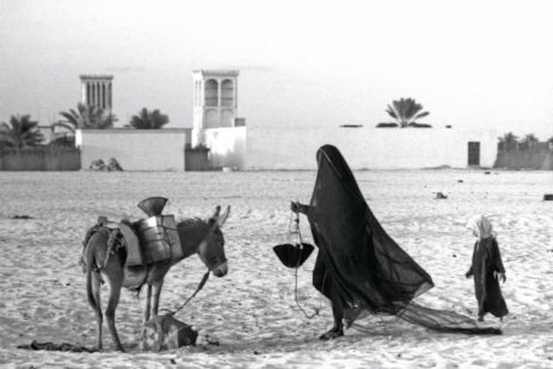 QASR AL HOSN HISTORY PROJECT 2013. Trucial Oman States, Abu Dhabi. An photograph of Abu Dhabi taken by Ronald Codrai. Image shows a woman collecting water fom outside the fort. in the background are wind towers. 

Ronald Codrai © TCA Abu Dhabi