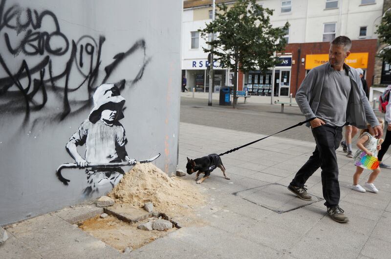 A man and his dog walk past an image of a girl making a sandcastle with a crowbar in Lowestoft, a mural very much in the style of Banksy.