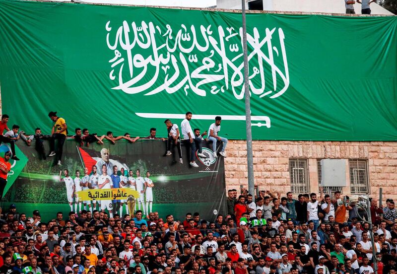 Football fans stand beneath a giant banner depicting the Saudi national flag and a smaller one reading in Arabic "Long live the Fedayeen (Palestinian national football team nickname)" and showing Palestine's players and coach Noureddine Ould Ali (C), as they attend the World Cup 2022 Asian qualifying match between Palestine and Saudi Arabia in the town of al-Ram in the Israeli occupied West Bank. The game would mark a change in policy for Saudi Arabia, which has previously played matches against Palestine in third countries. Arab clubs and national teams have historically refused to play in the West Bank, where the Palestinian national team plays, as it required them to apply for Israeli entry permits. AFP