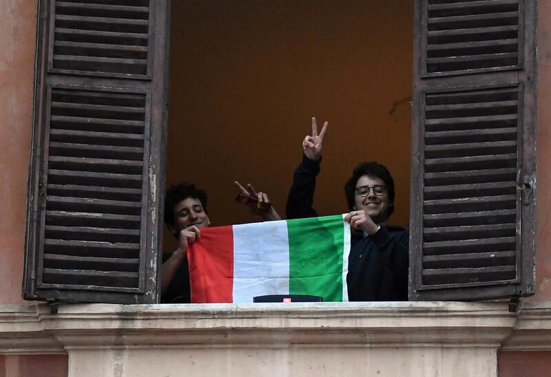 Men gesture holding an Italian flag as they look out of an apartment window as part of a flashmob organised to raise morale during Italy's coronavirus crisis in Rome, Italy, March 13, 2020. REUTERS/Alberto Lingria