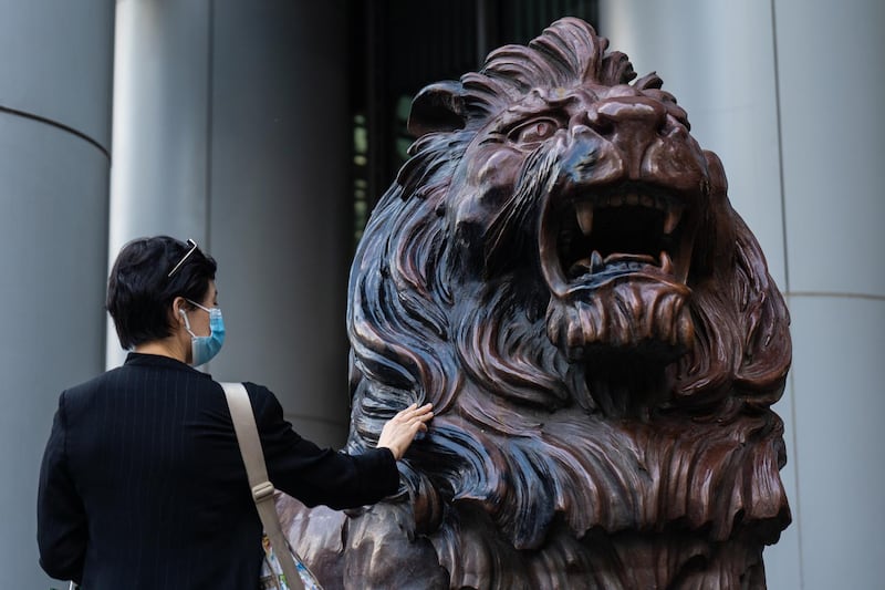 A person wearing a protective mask touches a statue of a lion in front of the HSBC Holdings Plc headquarters building in Hong Kong, China, on Thursday, Oct. 22, 2020. Damaged and defaced during tumultuous protests at the start of this year, the two bronze lion statues standing guard outside HSBC's main office in Hong Kong have made their return to the public in a city subdued by a Chinese crackdown on dissent. Photographer: Chan Long Hei/Bloomberg