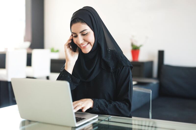 Cheerful middle eastern ethnicity woman is managing client calls and her private business at home using laptop and smartphone. She is talking to someone on the phone while checking information online. Dressed in black religious veil, has a wedding ring. Image contains copy space. Made in Dubai.