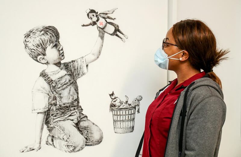 A handout picture recieved from University Hospital Southampton on May 7, 2020 shows a member of staff posing with an artwork by street artist Banksy called "Game Changer", showing a boy playing with a nurse superhero toy with figures of Batman and Spiderman discarded in a basket as a tribute to NHS staff who are continuing to work during the COVID-19 pandemic, on a wall at the University Hospital Southampton, southern England. - A new artwork by Banksy in honour of Britain's health service has gone on display in a hospital, paying tribute to medics battling the coronavirus pandemic in the second hardest-hit country. The street artist also posted an image of the work on Instagram, which shows a boy in dungarees playing with a figurine of a nurse in a superhero cape. (Photo by Stuart MARTIN / various sources / AFP) / RESTRICTED TO EDITORIAL USE - MANDATORY CREDIT "AFP PHOTO / UNIVERSITY HOSPITAL SOUTHAMPTON / STUART MARTIN " - NO MARKETING - NO ADVERTISING CAMPAIGNS - MANDATORY MENTION OF ARTIST UPON PUBLICATION - DISTRIBUTED AS A SERVICE TO CLIENTS