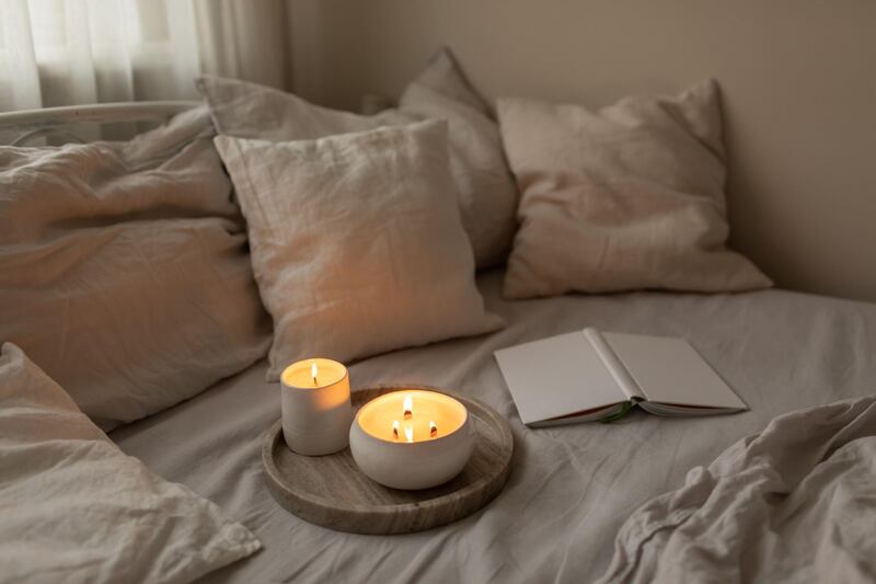 Organic soya wax candles with wooden candlewick in white ceramic bowls on linen bedding set with book. Cosy and soft winter background. Hygge and aromatherapy concept. Getty Images