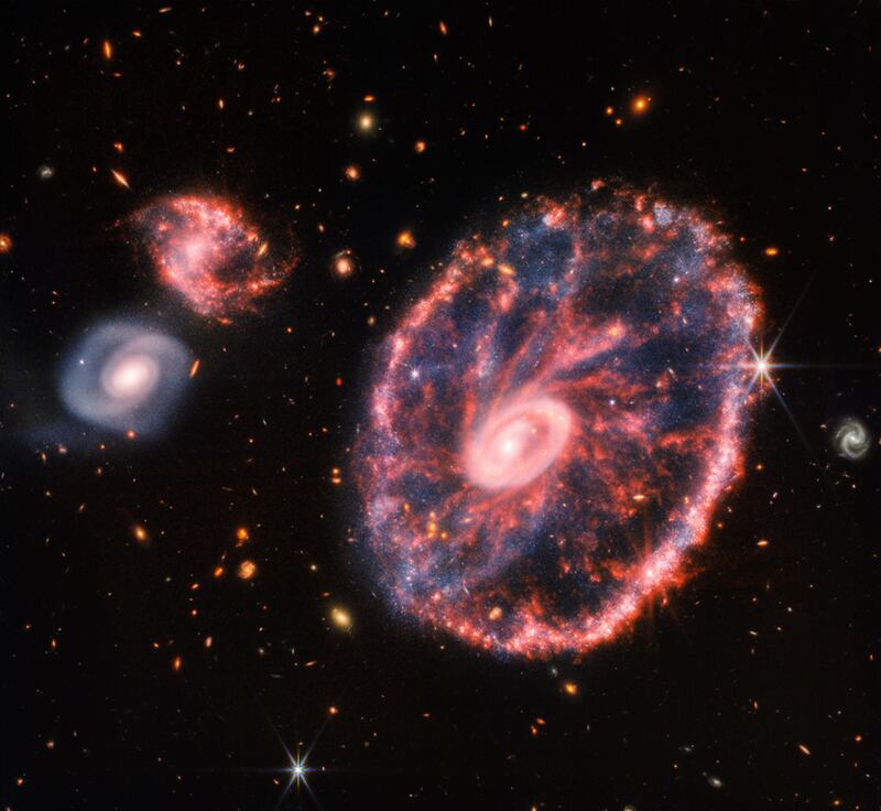 A large pink, speckled galaxy resembling a wheel with a small, inner oval, with two smaller spiral galaxies about the same size to the left, captured by the James Webb Space Telescope.  NASA