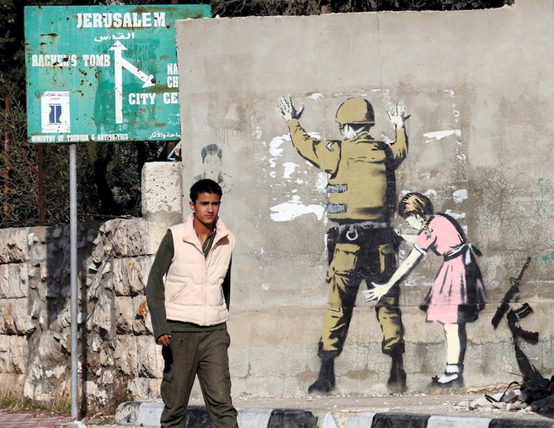 epa01191235 Illusive British graffiti artist named Banksy has painted new works in the West Bank town of Bethlehem, including a stencil work showing a small girl in a dress (a Palestinian?) frisking a soldier (an Israeli?) painted on a wall a Palestinian teenager passes by in the West bank town of Bethlehem, 04 December 2007. The works are not signed.  EPA/JIM HOLLANDER *** Local Caption *** 01191235