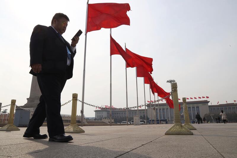A man uses his smartphone as he passes a row of red flags in Tiananmen Square near the Great Hall of the People where a session of the National People's Congress is held in Beijing, China, Friday, March 8, 2019. (AP Photo/Ng Han Guan)