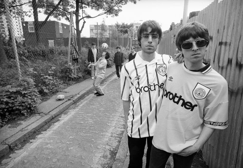 Singer Liam Gallagher and his guitarist brother Noel Gallagher of rock band Oasis, pose in Manchester City football shirts, 9th May 1994. Other band members can be seen in the background (left to right, from 2nd left) bassist Paul McGuigan, drummer Tony McCarroll and rhythm guitarist Paul 'Bonehead' Arthurs. (Photo by Kevin Cummins/Getty Images)  