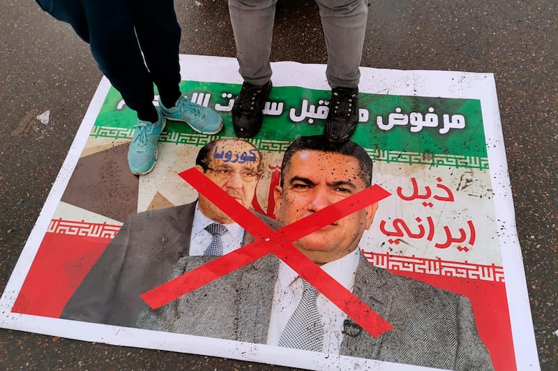 Anti-government protesters stand on a defaced poster with pictures of former Iraqi Prime Minister Nouri al-Maliki, left, and Iraq's Prime Minister-designate Adnan Al-Zurfi with an Arabic sentence reading "Rejected, Iranian tail, Iran followers" in Tahrir Square, Baghdad, Iraq, Wednesday, March 18, 2020. Iraq's president named Al-Zurfi as prime minister-designate, following weeks of political infighting and a looming crisis amid a global pandemic. (AP Photo/Hadi Mizban)