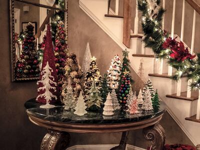 Decorative lighted Christmas trees decorate table with lighted garland on stair railing in background.