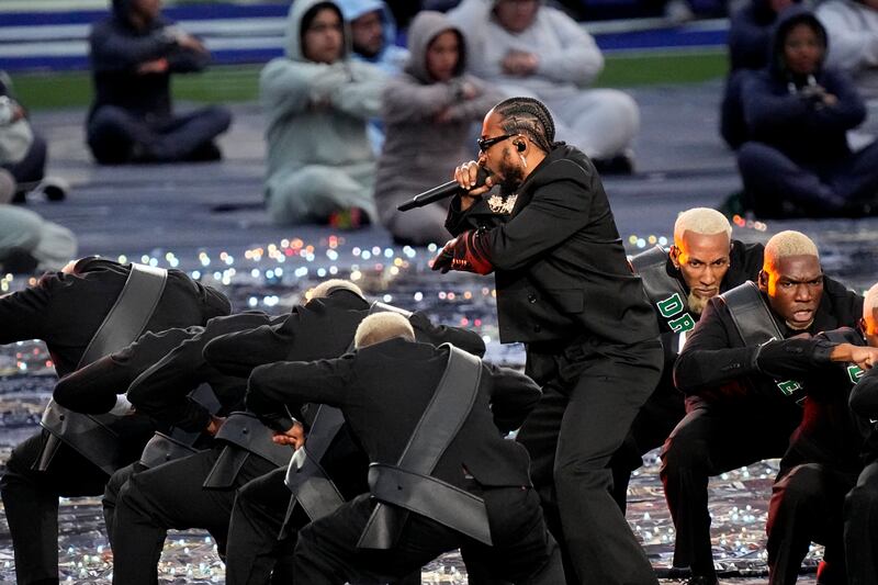 A black suit-wearing Kendrick Lamar performs alongside other men sporting the same outfit as him. AP