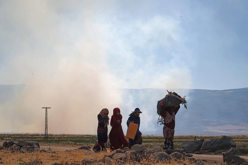 Syrian women walk by as smoke billows in the area of the Syrian village of az-Ziyarah, in the agricultural al-Ghab plain of the Hama province, on June 6, 2021, following reported pro-government bombardment. / AFP / Abdulaziz KETAZ
