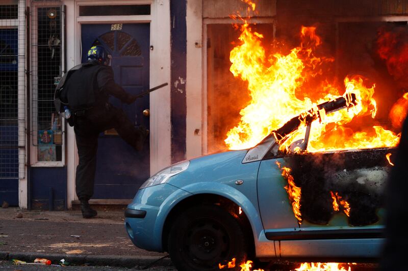 A riot police officer attempts to break down the door of a house next to a burning car in Hackney.