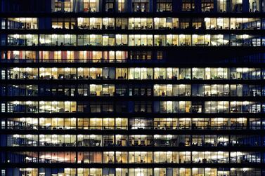 Lots of people working late. Employees seen as silhouettes against their brightly lit offices with large windows. Building lit by the "blue hour" evening sky. Getty Images