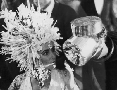 Elizabeth Taylor in a headdress of silver spikes, orchids and lilies chats to designer Pierre Cardin who is wearing what looks like an aluminium gas mask.   (Photo by Three Lions/Getty Images)