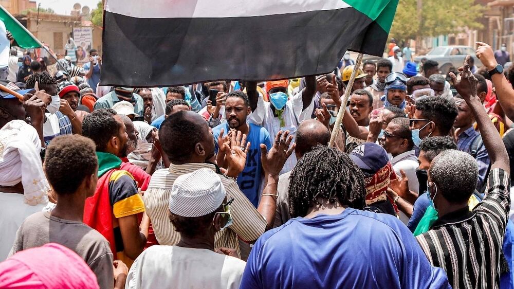 An anti-coup protester waves a Sudanese national flag while others chant slogans during mass demonstrations against military rule in the centre of Sudan's capital Khartoum on June 30, 2022.  - At least six Sudanese demonstrators were killed as security forces sought to quash mass rallies of protesters demanding an end to military rule, pro-democracy medics said.  In one of the most violent days this year in an ongoing crackdown on the anti-coup movement, AFP correspondents reported security forces firing tear gas and stun grenades to disperse tens of thousands of protesters.  (Photo by AFP)