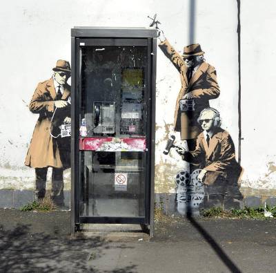 epaselect epa04167836 A general view of a payphone where a new mural believed to be by graffiti artist 'Banksy' was discovered in a residential street in Cheltenham, Britain, 15 April 2014. The painting showing intelligence agents engaged in wiretapping appeared next to a phone booth over the weekend and has yet to be authenticated. The new artwork comes in the wake of the storm over surveillance by British intelligence agency GCHQ and US intelligence agency NSA revealed by US whistleblower Edward Snowden. GCHQ is also based in Cheltenham.  EPA/NEIL MUNNS