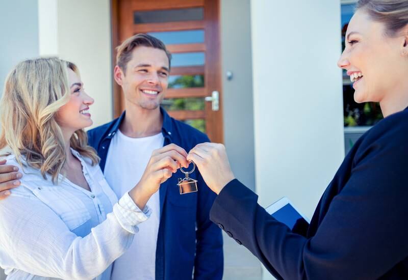 Real estate agent giving a young couple the key to their new house. The house is contemporary. All are happy and smiling. The couple are casually dressed and the agent is in a suit. The front door is also visible. Copy space