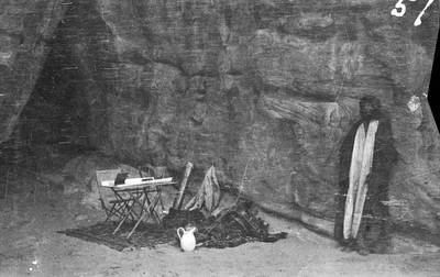 Shakespear’s camping gear and Arab figure by rock face and cave, probably in mountains in vicinity of Wadi Iram/Yatun Aqabah.
