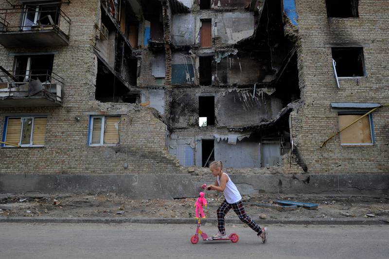 A girl rides a scooter past a destroyed building in the village of Horenka, Kyiv. AFP