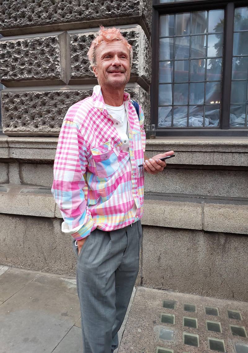 A man wears a pink checked shirt with grey trousers. Extra points for the pink hair. 